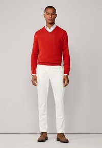 Punainen V-kaula-aukkoinen neule valkoisen kauluspaidan päällä, yhdistettynä valkoisiin housuihin ja ruskeisiin nahkasaappaisiin. Sileä kangas, istuva malli, siistit linjat.