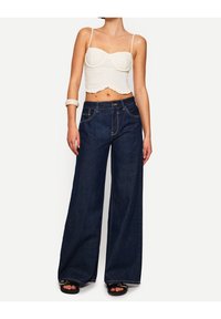 Tmavě modré džíny s širokými nohavicemi a kontrastním prošitím, s předními kapsami a vysokým pasem. V kombinaci s bílým zkráceným topem a černými sandály.