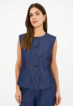 Donna con lunghi capelli castani che indossa un top blu senza maniche con bottoni e pantaloni abbinati, in piedi su uno sfondo bianco uniforme.