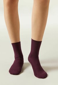 Piernas con calcetines oscuros morados de canalé hasta los tobillos, de pie sobre un fondo claro y neutral.