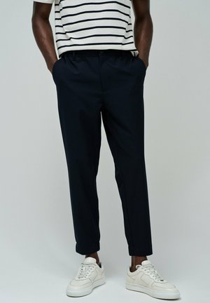 Pantalones azul marino oscuros con un corte ajustado, que presentan una textura suave y bolsillos laterales, combinados con zapatillas blancas y una camisa a rayas.