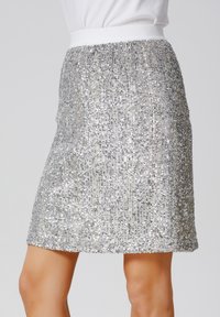 Jupe en sequins argentés avec une ceinture élastique blanche ajustée, longueur genou, présentant une surface texturée et un éclat subtil des sequins.