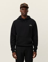 Czarna bluza z kapturem, ma kieszeń z przodu i sznurek do regulacji, z małym białym logo na klatce piersiowej. Nosi się ją z czarnymi spodniami i czapką w kratę.