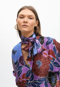 Blusa a maniche lunghe con stampa floreale viola, dotata di un nodo al collo, con accenti blu, borgogna e verdi. Tessuto morbido con un design vivido.