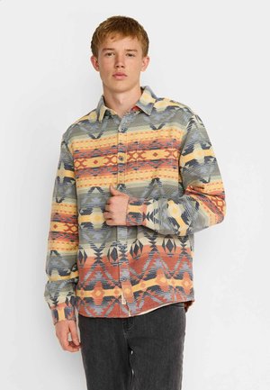 Joven con el pelo corto y rojo, vistiendo una camisa abotonada con un patrón geométrico multicolor y vaqueros oscuros, de pie sobre un fondo blanco liso.