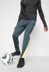 Atletické kalhoty Nike v tmavě tyrkysové barvě s žlutým akcentem. Vyrobené z hladkého materiálu, s přiléhavým střihem, v kombinaci s černými fotbalovými botami a míčem.