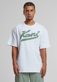 White cotton t-shirt with short sleeves, featuring green textured "Kani" logo on the front. Loose fit, round neckline.
