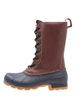 Botte imperméable en cuir brun et caoutchouc noir avec laçage à l'avant, boucle de traction à l'arrière, et semelle rainurée beige pour une bonne adhérence.