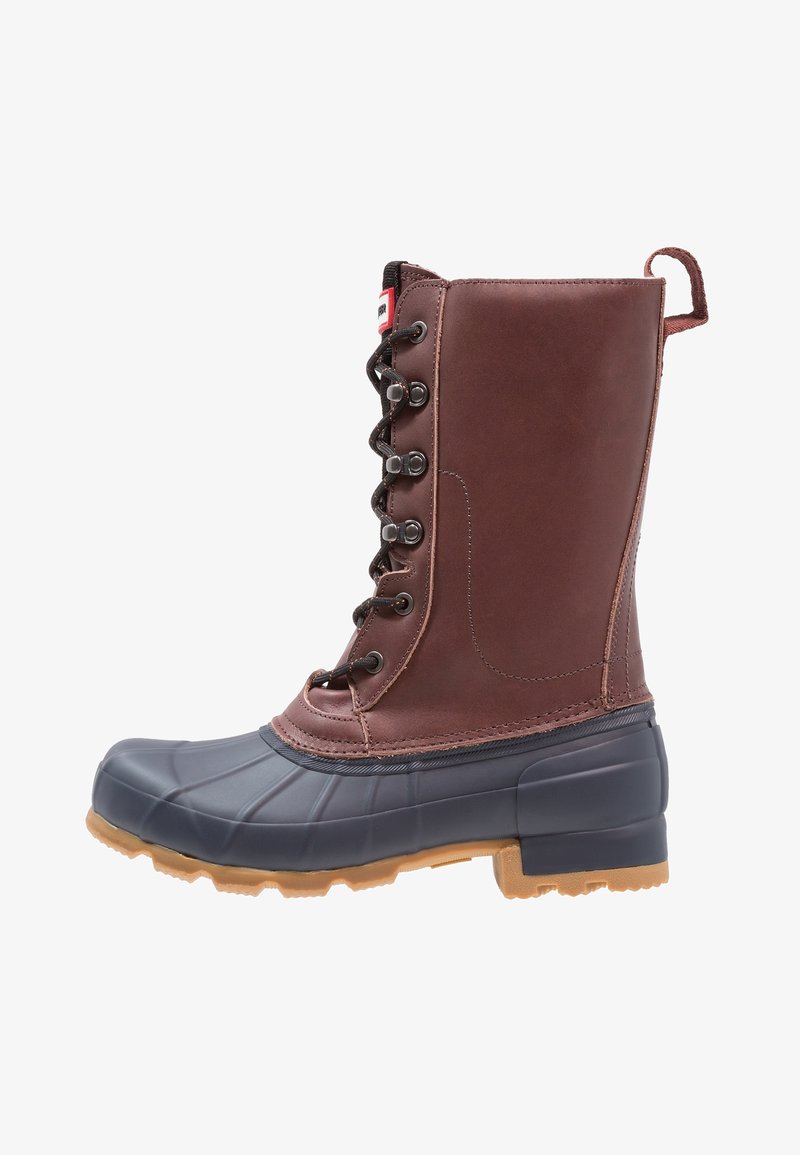 Botte imperméable en cuir brun et caoutchouc noir avec laçage à l'avant, boucle de traction à l'arrière, et semelle rainurée beige pour une bonne adhérence.