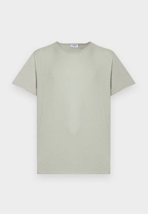 Młody mężczyzna z kręconymi włosami, ubrany w jasnooliwianą koszulkę, pasujące szorty i czarne sandały, stoi z rękami w kieszeniach.