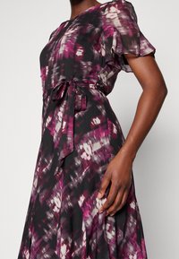 Svart klänning med ett rosa och vitt abstrakt mönster, korta svävande ärmar, midjeband och flytande kjol. Tillverkad av lättviktigt tyg.