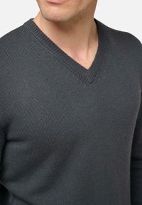 Homme portant un pull en maille gris foncé à col en V, montrant le décolleté et le haut de la poitrine, devant un fond clair uni.