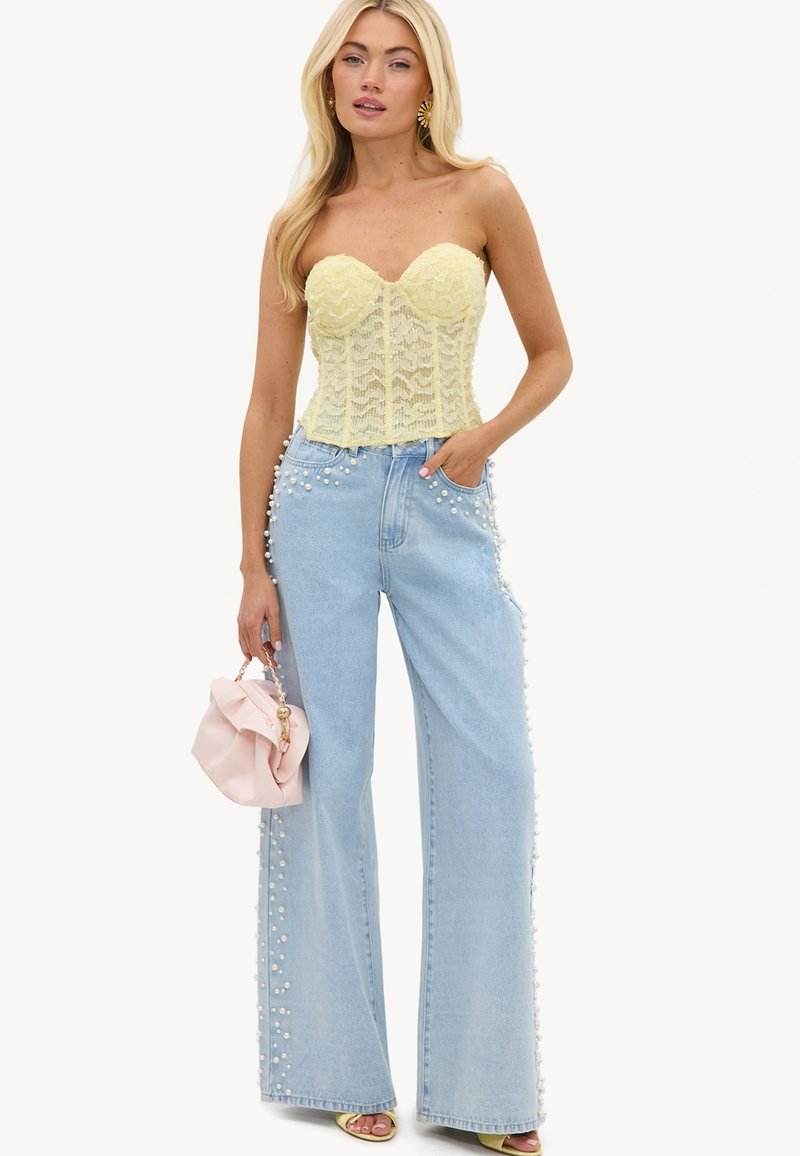 Femme portant un corset jaune en dentelle sans bretelles et un jean bleu clair à jambes larges décoré de perles, tenant un sac à main rose avec chaîne dorée.