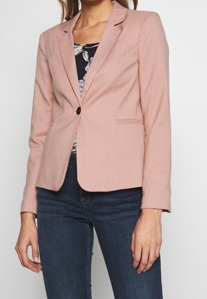 Vrouw draagt een nauwsluitend stofroze colbert met een enkele zwarte knoop, een bloemenprint top en donkerblauwe spijkerbroek tegen een effen witte achtergrond.