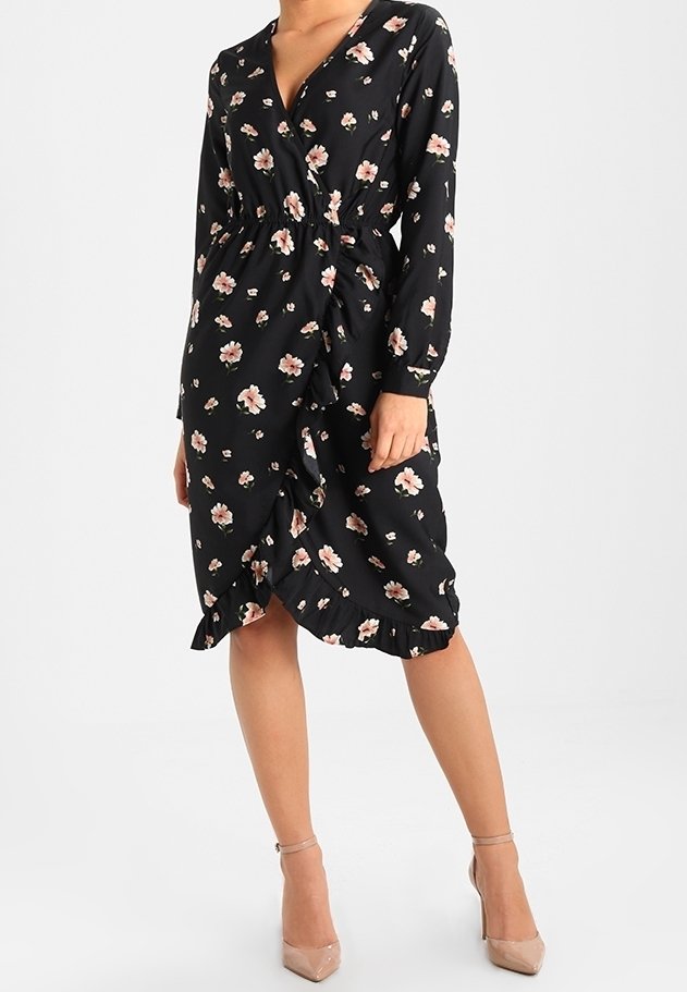 Femme portant une robe portefeuille noire à imprimé floral rose, manches longues, détails à volants et escarpins nude à bride cheville.