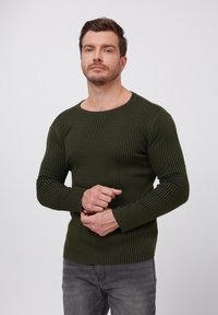Suéter de punto acanalado verde oscuro con cuello redondo, diseño ajustado y mangas largas, combinado con jeans grises. Patrón texturado visible en toda la prenda.