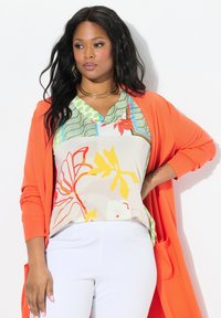 Leuchtend orangefarbener langer Cardigan über einem mehrfarbigen Blusenshirt mit floralen Mustern in Grün, Rot und Gelb. Kombiniert mit hellen weißen Hosen.