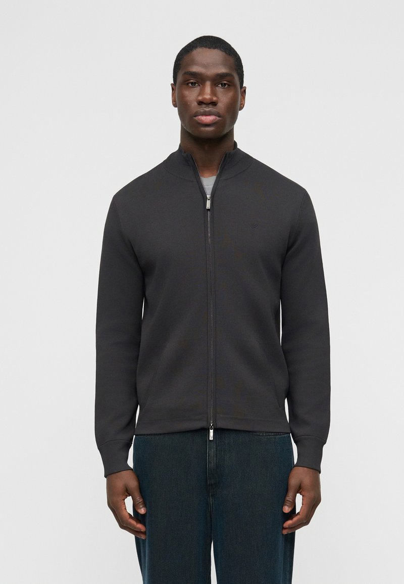 Νέος άνδρας που φοράει μαύρο μπουφ άν τύπου bomber με φερμουάρ και σκουρόχρωμο μπλε τζιν, στέκεται μπροστά σε απλό λευκό φόντο.