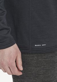 Mørkegrå hurtigtørrende langærmet skjorte med tekstureret stof, flade sømme og et label med "QUICK DRY" i hvidt nær sømmen.
