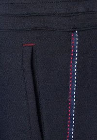 Černá atletická tkanina s texturovaným povrchem. Obsahuje viditelný červený steh vedle bílých akcentů a přední otvor kapsy.