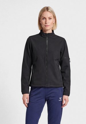 Mujer con chaqueta negra con cremallera y pantalones deportivos azules, de pie frente a un fondo blanco liso.