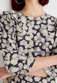 Pukupuku mekko teksturoidusta kankaasta, jossa on laivastonsininen ja beige leopardikuosi. Kolme neljännesosaa hihat, joissa on pleat-tyylinen muotoilu ja hienovaraiset napin yksityiskohdat.
