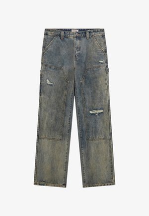 Chemise vert olive à manches courtes portée sur un haut noir à manches longues, associée à un jean ample bleu clair déchiré et des baskets noires.