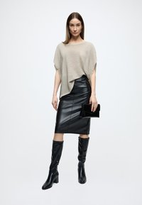 Top bej tricotat supradimensionat, fustă creion neagră din piele ecologică, cizme negre până la genunchi, ținând o poșetă clutch neagră. Texturi fine, siluetă ajustată.