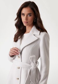 Manteau blanc en mélange de laine avec une coupe ajustée, grands revers, boutons dorés et ceinture nouée à la taille. Texture lisse avec des coutures structurées.