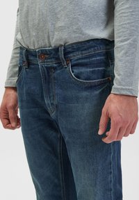 Ciemnoniebieskie dżinsy z klasycznym designem pięciu kieszeni, z zapięciem na guzik i jasnymi detalami przeszycia. Widoczna tekstura w stanie niepranym.