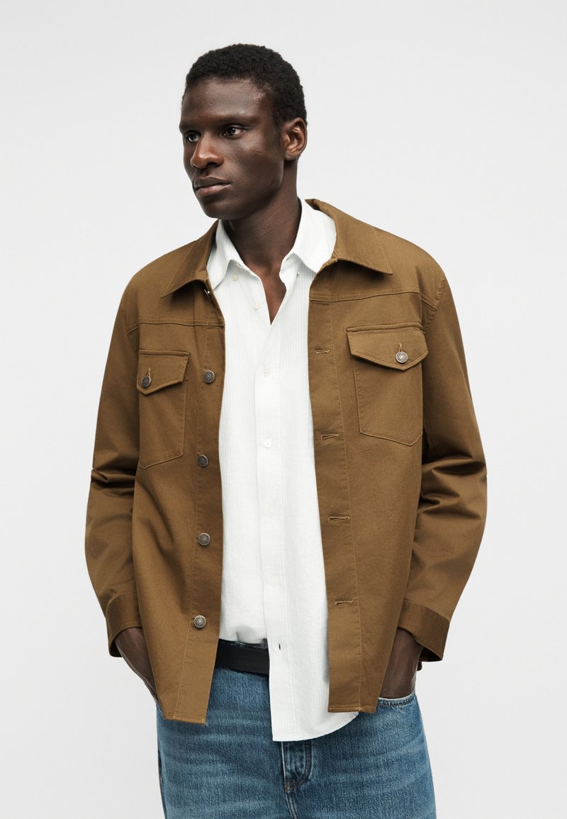 Mann trägt eine braune, geknöpfte Jacke über einem weißen Hemd und blauen Jeans, steht mit den Händen in den Taschen und schaut nach links vor neutralem Hintergrund.