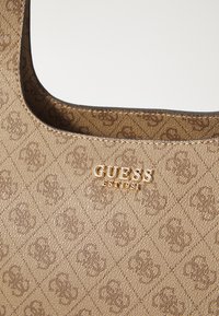 Bruine handtas met een patroon, gekenmerkt door een gladde textuur, ronde vorm en een gouden logo met "GUESS EST 1981" dat prominent is weergegeven.