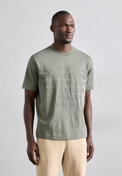 T-shirt in cotone verde oliva con collo a giro, maniche corte e un design a logo in rilievo tono su tono sul davanti. Vestibilità regolare, texture liscia.