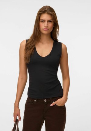 Young woman with long brown hair wearing a black sleeveless V-neck top and dark brown pants holding a brown handbag with one hand in pocket.
