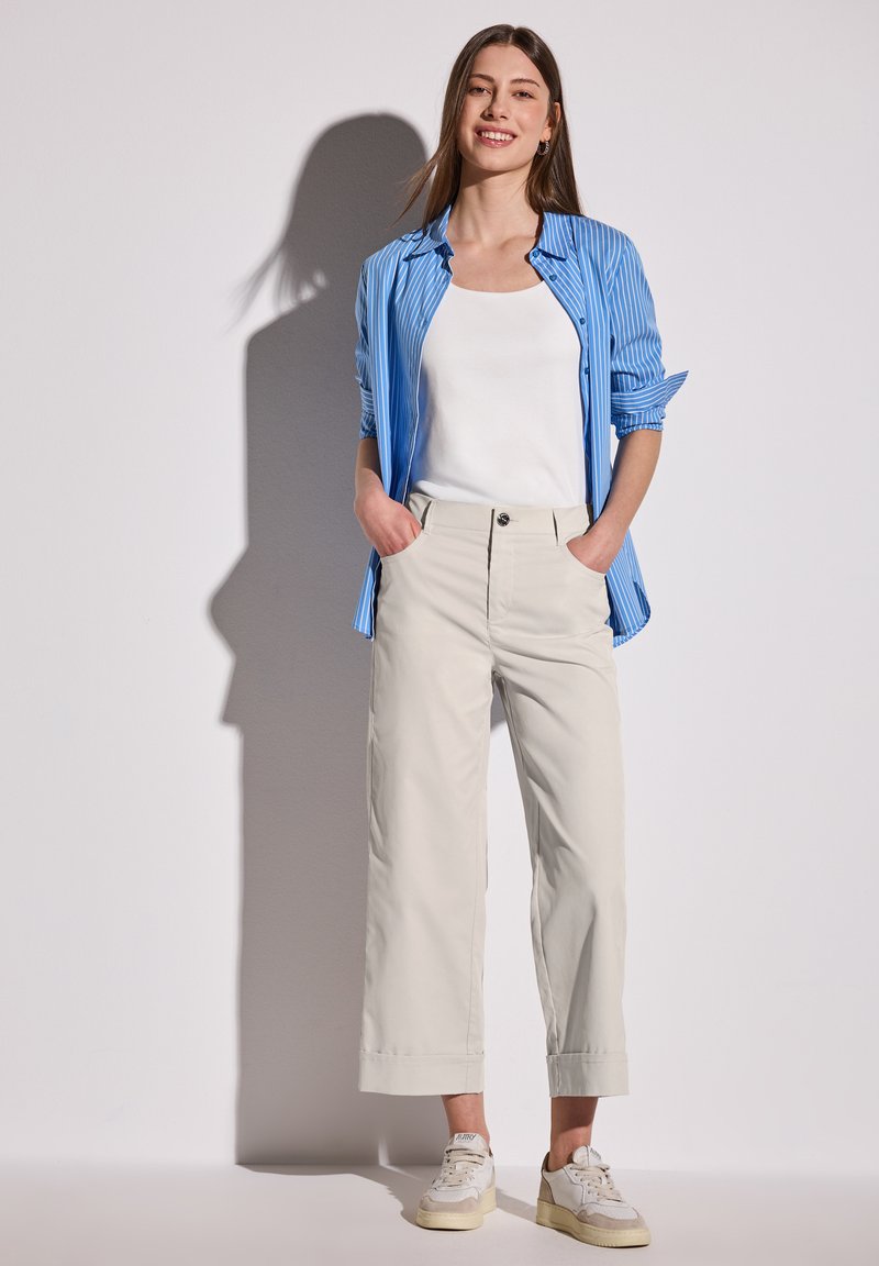 Ljusbeiga vida byxor med hög midja, kombinerade med en blårandig skjorta och en vit ärmlös topp. Casual sneakers fulländar looken.