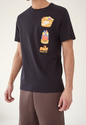 Homme portant un t-shirt Nike noir avec un panier de fruits colorés et des graphiques de sac à provisions, assorti à un pantalon marron.