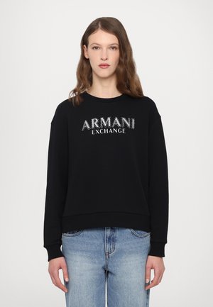 Zwarte sweatshirt met "ARMANI EXCHANGE" in glanzend zilveren letters, ronde hals, lange mouwen en ribgebreide manchetten, gecombineerd met lichtblauwe jeans.