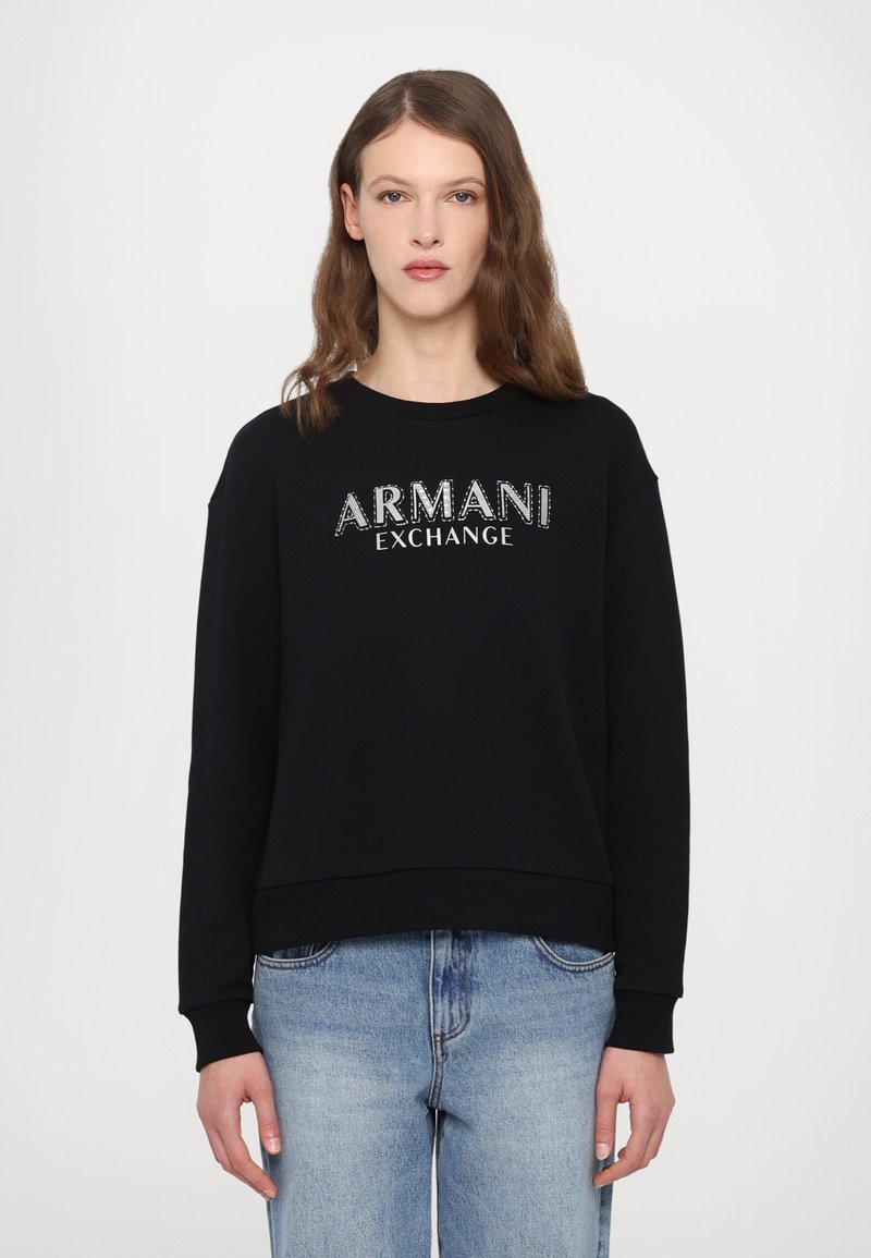 Zwarte sweatshirt met "ARMANI EXCHANGE" in glanzend zilveren letters, ronde hals, lange mouwen en ribgebreide manchetten, gecombineerd met lichtblauwe jeans.