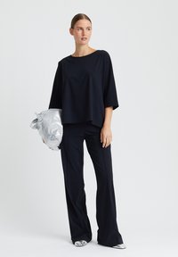 Schwarze weit geschnittene Hose und lockeres Oberteil, beide aus glattem Stoff. Das Modell hält eine silberne Tasche. Die Schuhe haben metallische Akzente. Sauberes, minimalistisches Design.