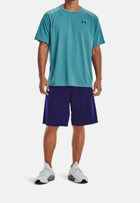 Męski strój sportowy składający się z turkusowej koszulki odprowadzającej wilgoć i fioletowych spodenek, uzupełniony białymi sneakersami z czarnymi akcentami.