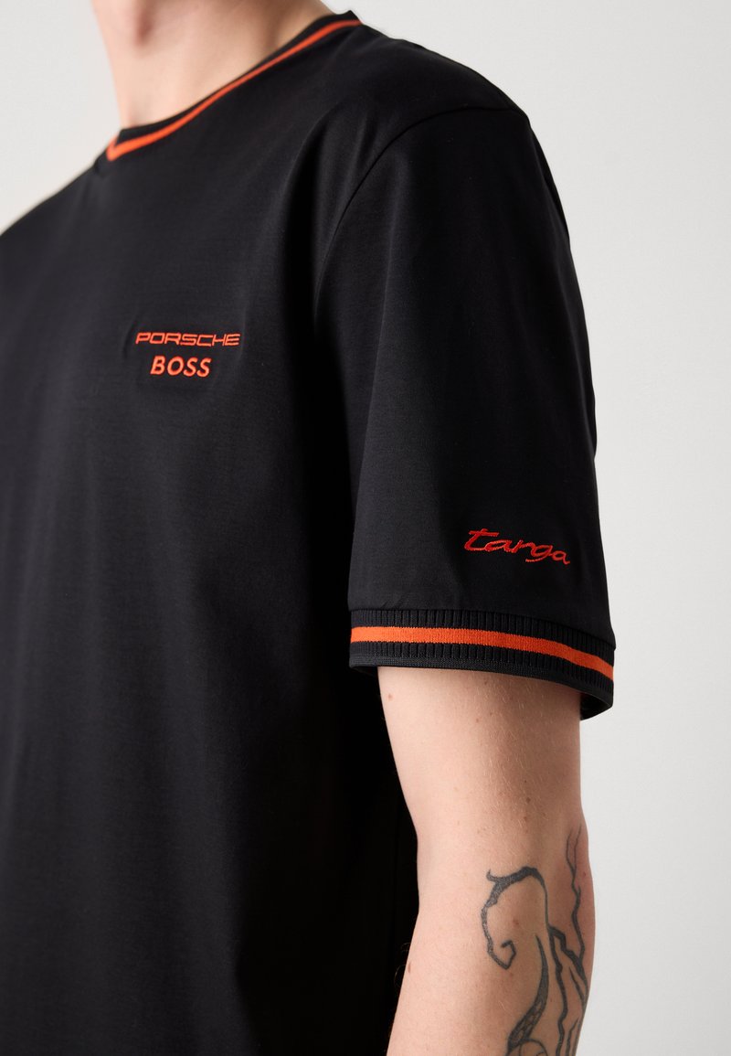 T-shirt noir à manches courtes, avec un col et des accents de manches côtelés orange. Logos "Porsche" et "BOSS" brodés.