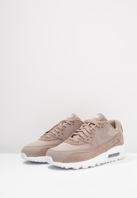 Tenisky Nike Air Max 90 mají světle hnědou svršek z semiše a síťoviny, bílou podrážku, kulatou špičku a zavazování na tkaničky. Boční pohled odhaluje vzduchové polstrování.