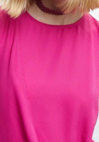 Blouse fuchsia à texture lisse, avec un col rond et un détail plissé subtil à l'avant. Collier ras de cou perlé.