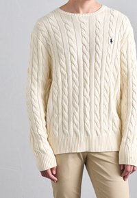 Krémový svetr s kabelovým pletením, s žebrovaným lemem a manžetami, s malým vyšitým logem na levé hrudi. Vyrobeno z měkkého, jemně strukturovaného materiálu.