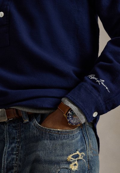 Camisa de manga longa azul marinho com punhos abotoados, apresentando um forro interior cinza e logótipo bordado. Usada com jeans desgastados e um relógio.