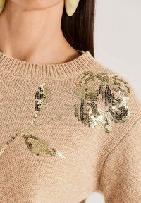 Frau in beigem Strickpullover mit goldfarbener, bestickter Blumenverzierung an der Schulter und großen, hellgelben Ohrringen.