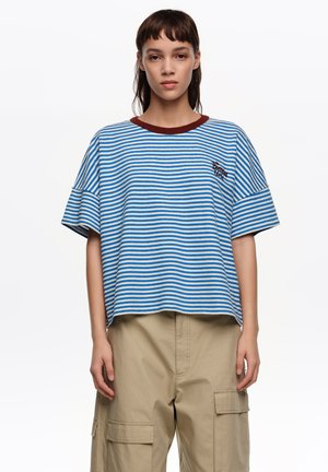 Joven con camiseta de manga corta a rayas azules y blancas con cuello marrón y pantalones cargo beige, posando frente a un fondo blanco.