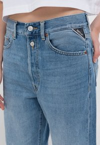 Ljust blå denimjeans av medelhöjd med dubbla metallknappar, framfickor och en liten märkespatch på höger ficka, bärs av en person.