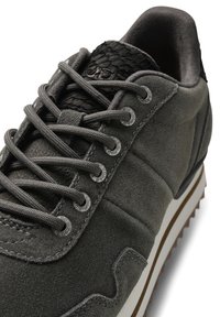 Gray suede sneaker featuring flat gray laces, a textured black leather accent at the heel, and a white rubber sole with a brown tread.