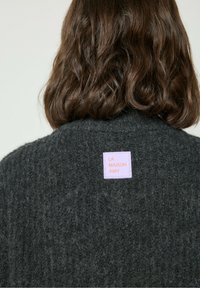 Pull en maille texturée gris foncé avec une étiquette rose "LA MAISON AMV" cousue à l'arrière. Le matériau semble doux et épais.